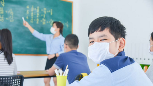 A boy wearing a mask looking at a camera in a classroom