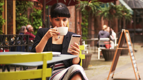 Zu sehen ist eine Frau, die in einem Straßenkaffee sitzt, einen Kaffee trinkt und eine Nachricht auf ihrem Smartphone liest.