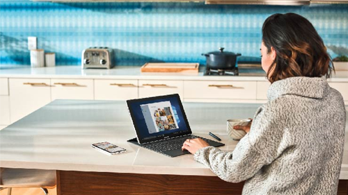 Zu sehen ist eine Frau an einem Küchentisch auf dem ein Surface steht und ein Smartphone daneben liegt.