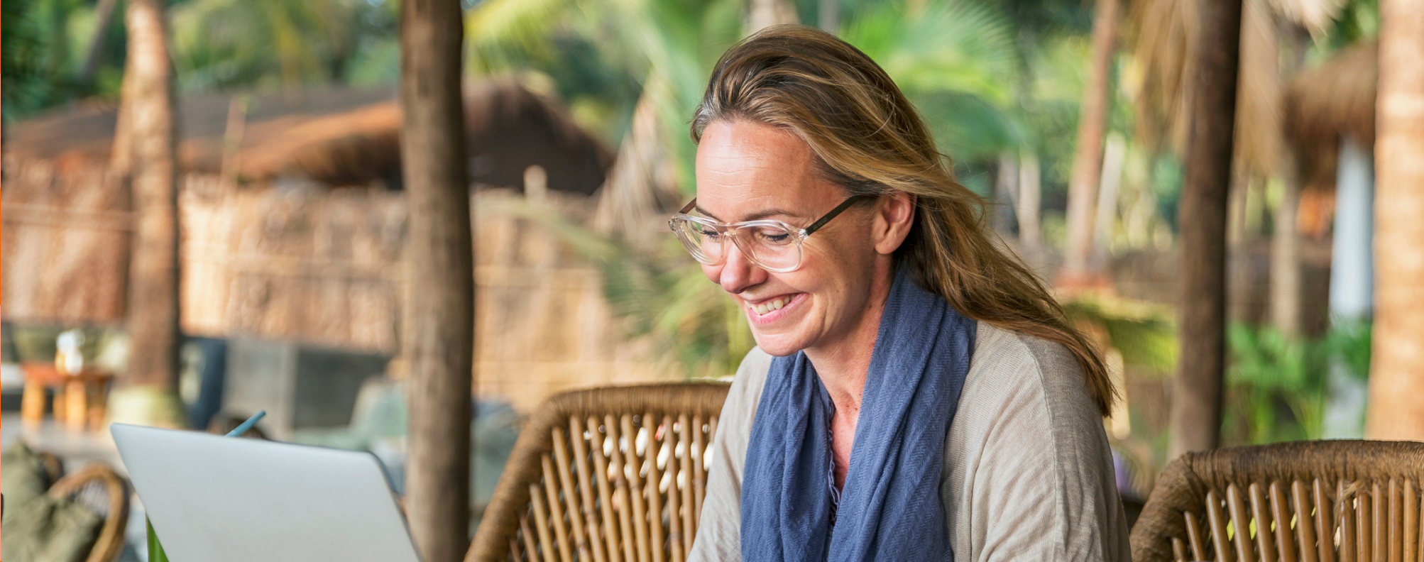 Person working on a laptop at an outdoor wooden table with tropical plants and thatched structures in the background.