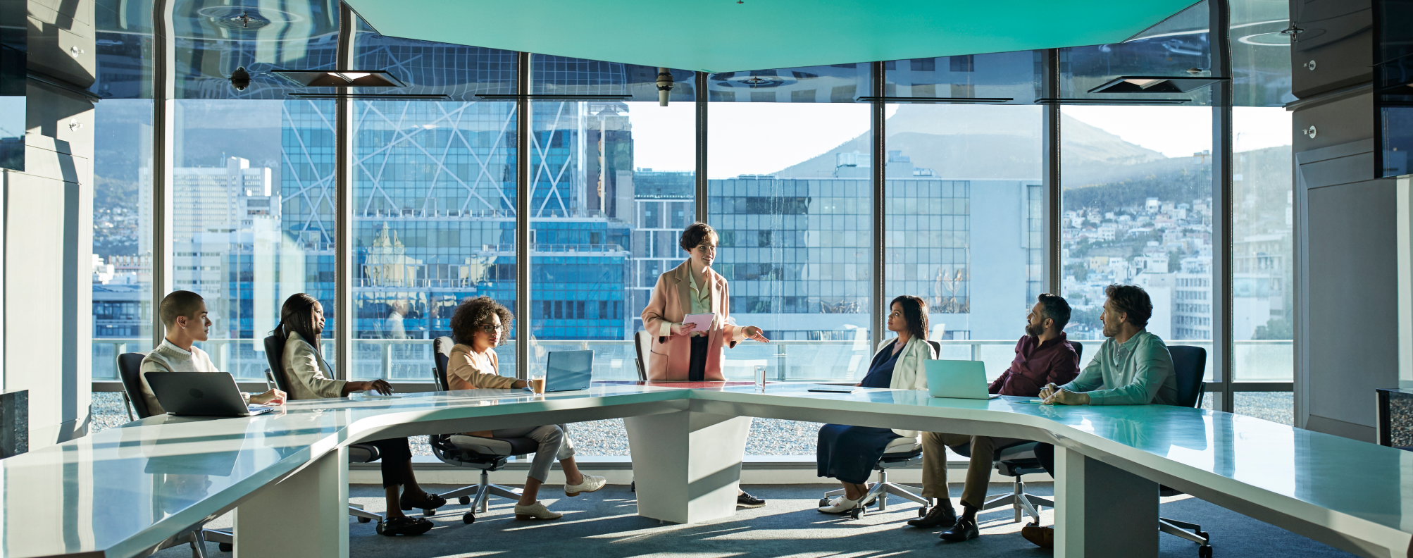 Seven people in a modern office meeting room with city and mountain views, one person presenting.