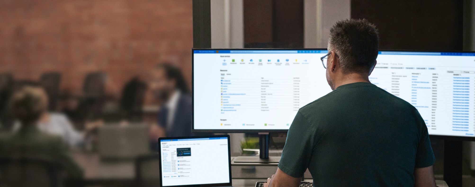 Person working at a desk with three monitors displaying file management software, viewed from behind. The background shows a blurred meeting room with people seated at a table.
