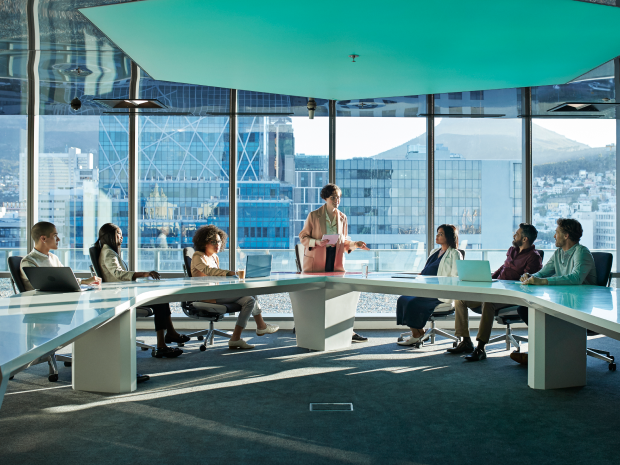 Seven people in a modern office meeting room with city and mountain views, one person presenting.
