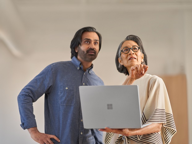 A man and a woman standing and collaborating in an office space. The woman is holding a laptop and they are both looking up at a screen out of frame.