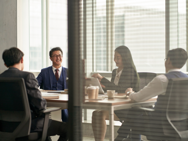  Four people in business attire seated around a conference table with notebooks, coffee cups, and documents in a modern office setting.