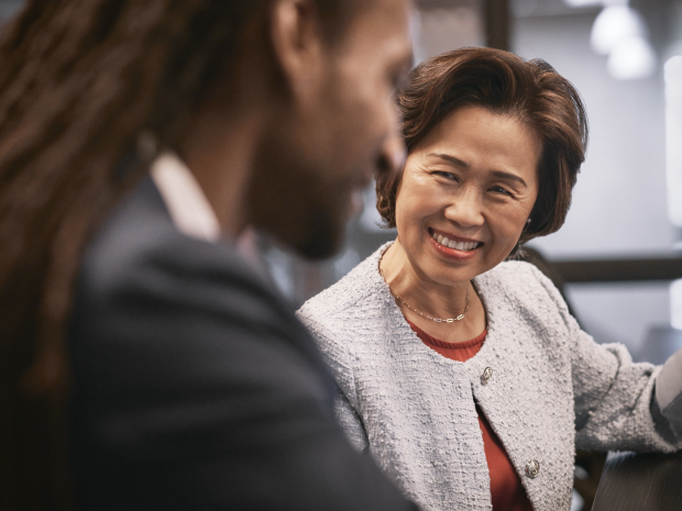Two professionals seated in conversation at a modern office table, with focus on one person’s styled hair, necklace, and textured jacket against a softly lit background.