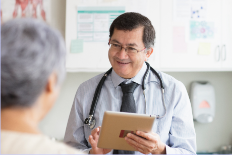 A smiling doctor holding a tablet and talking to a patient.