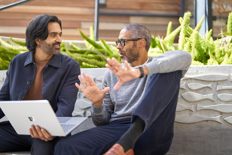 Two people sitting outside talking while one holds a laptop