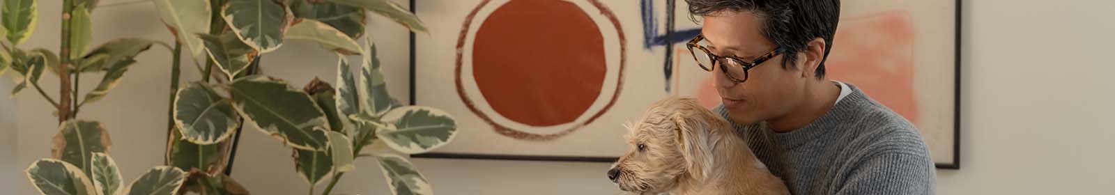 A man sits at a table with a dog on his lap and looks at a laptop