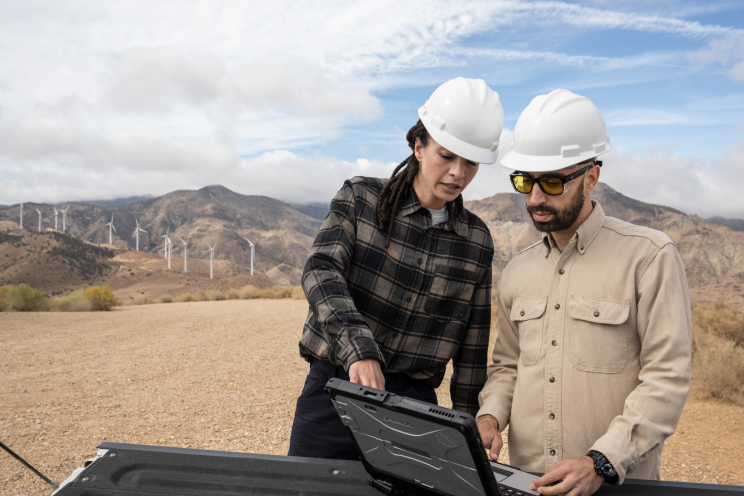 Two people in hardhats at a wind farm looking at a laptop