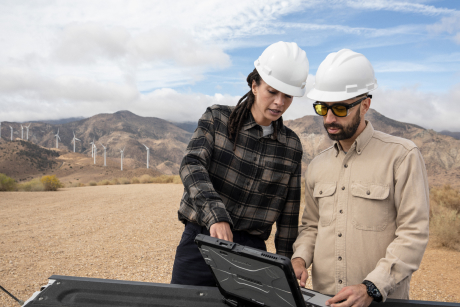 Two people in hardhats at a wind farm looking at a laptop