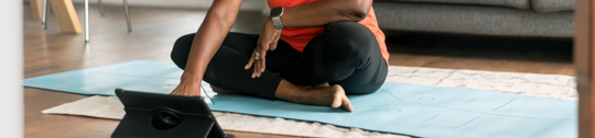 A senior woman participating in an online yoga class at home, interacting with tablet while sitting on a blue yoga mat