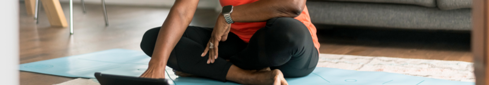A senior woman participating in an online yoga class at home, interacting with tablet while sitting on a blue yoga mat