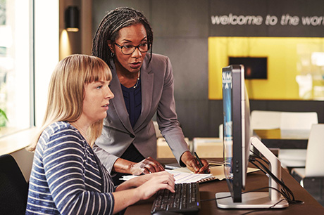 Two women at a desk look at a computer screen One of the women is standing while the other is seated and using a keyboard