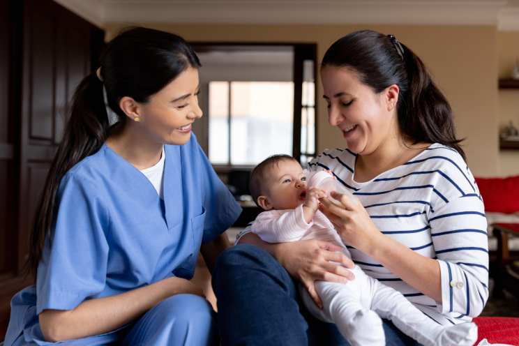 A person giving a bottle to a baby supervised by a nurse.