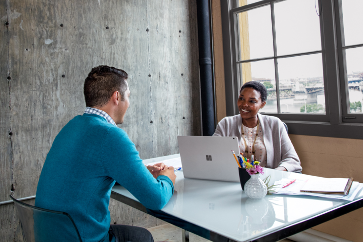 Two people seated at a desk talking over a laptop