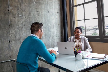 Two people seated at a desk talking over a laptop