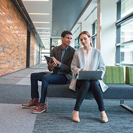 Two coworkers sitting on bench and working on devices