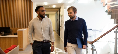 Two male professionals talk as they walk down a hallway