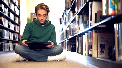 Small child sitting, reading a tablet in a library aisle