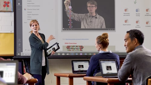 A teacher lectures at the front of a classroom with a tablet