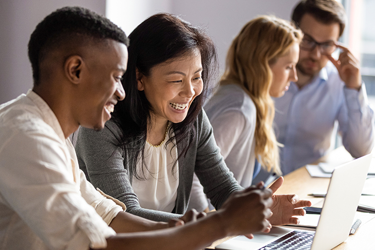 Four people sitting at a desk and smiling at a laptop