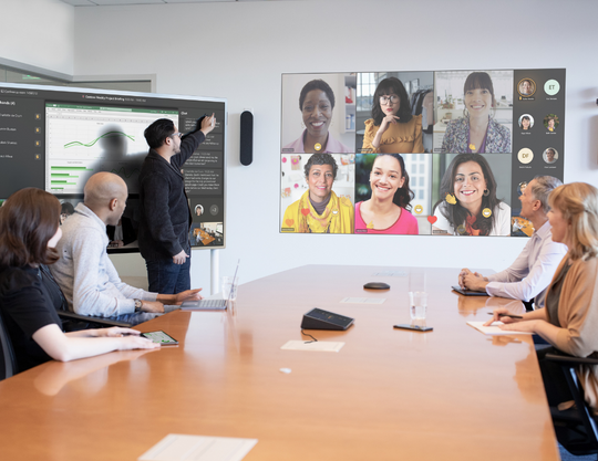 Four people stood around a table in a meeting, joined by virtual guests on a large screen
