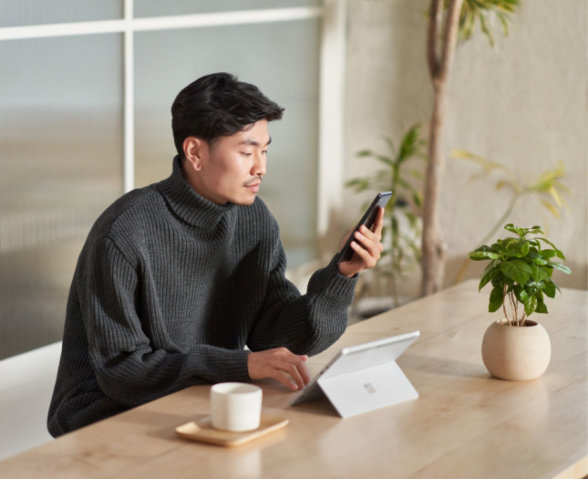 A man holding a mobile in his hand in in front of a tablet and a cup of coffee in a cafeteria