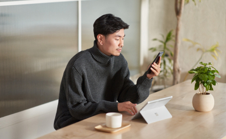 A man holding a mobile in his hand in in front of a tablet and a cup of coffee in a cafeteria