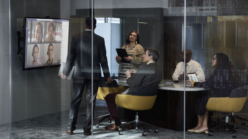 Ein Glaskonferenzraum mit einem Tisch um den 5 Personen sitzen und stehen. An der Wand ist ein Bildschirm mit  einem Microsoft Teams Meeting.