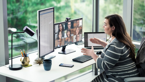 Eine Frau ist an einem Schreibtisch. Vor ihr sind zwei Bildschirme, ein Laptop mit Tastatur und Maus. Sie ist gerade in einer Videokonferenz.