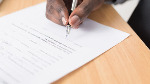 Es ist ein Vertrag auf einem Tisch zu sehen und eine Hand mit einem Stift.