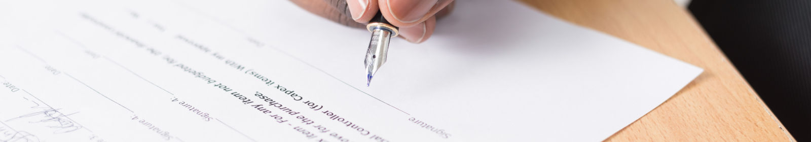 Es ist ein Vertrag auf einem Tisch zu sehen und eine Hand mit einem Stift.