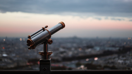 Man sieht ein Fernrohr im Vordergrund. Im Hintergrund ist eine Stadt bei der Dämmerung zu erkennen.
