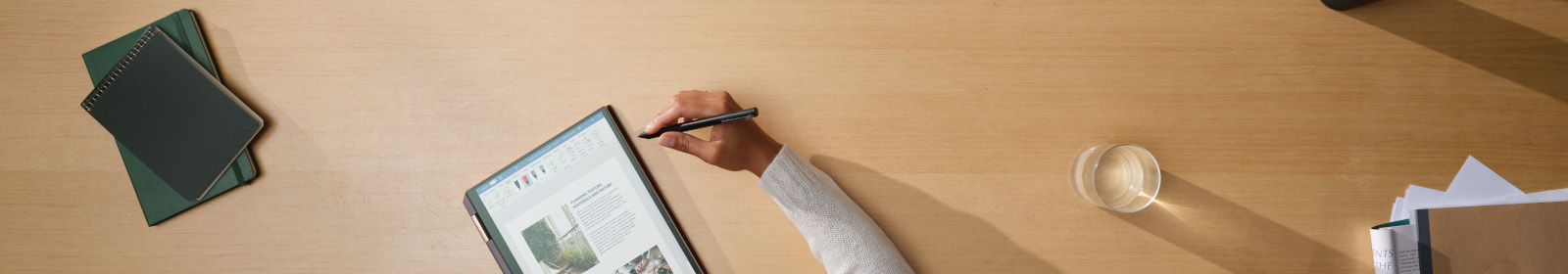 Man sieht einen Schreibtisch. Es liegen Bücher sowie Notizen auf dem Schreibtisch. In der Mitte ist ein Tablet zu sehen und eine Person arbeitet daran.