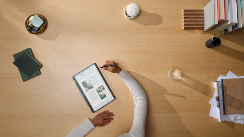 Man sieht einen Schreibtisch. Es liegen Bücher sowie Notizen auf dem Schreibtisch. In der Mitte ist ein Tablet zu sehen und eine Person arbeitet daran.