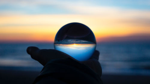 Im Hintergrund ist ein Sonnenuntergang am Meer. Im Vordergrund hält eine Hand eine Glaskugel.