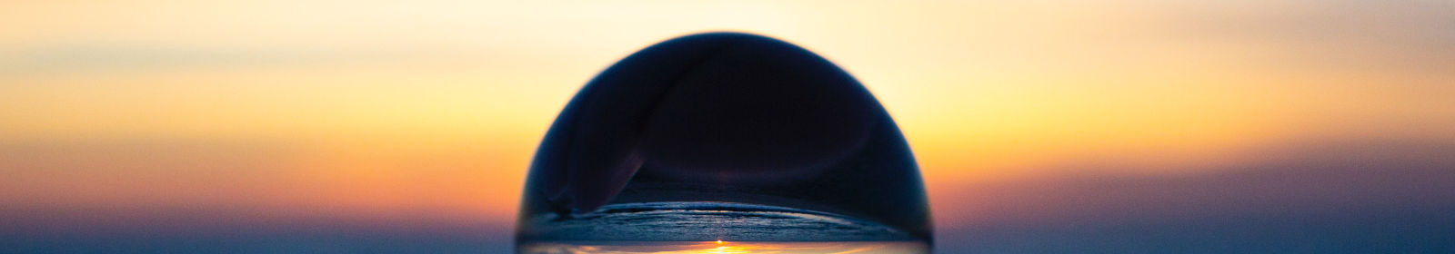 Im Hintergrund ist ein Sonnenuntergang am Meer. Im Vordergrund hält eine Hand eine Glaskugel.