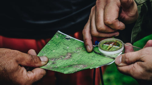 Im Vordergrund ist eine Landkarte und ein Kompass. Auf diesen zeigt eine Hand. Im Hintergrund sind zwei Menschen zu erkennen.