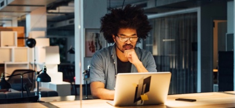 Man sitting at a desk in dimly lit room looking at laptop