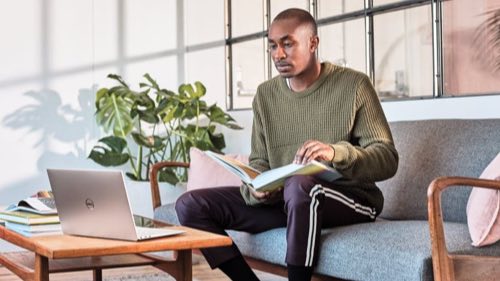 Person sitting on a couch with a book on his lap and a laptop on a coffee table