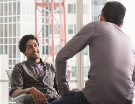 Two men talk with a cityscape behind them