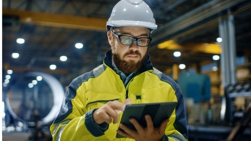 Man with beard and wearing a hard hat using a tablet in a factory like setting