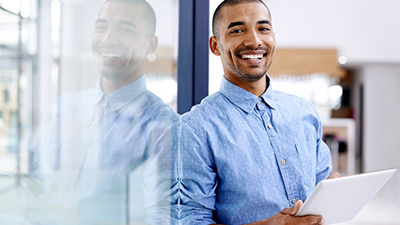 Male leaning on glass wall working on tablet and smiling at the camera